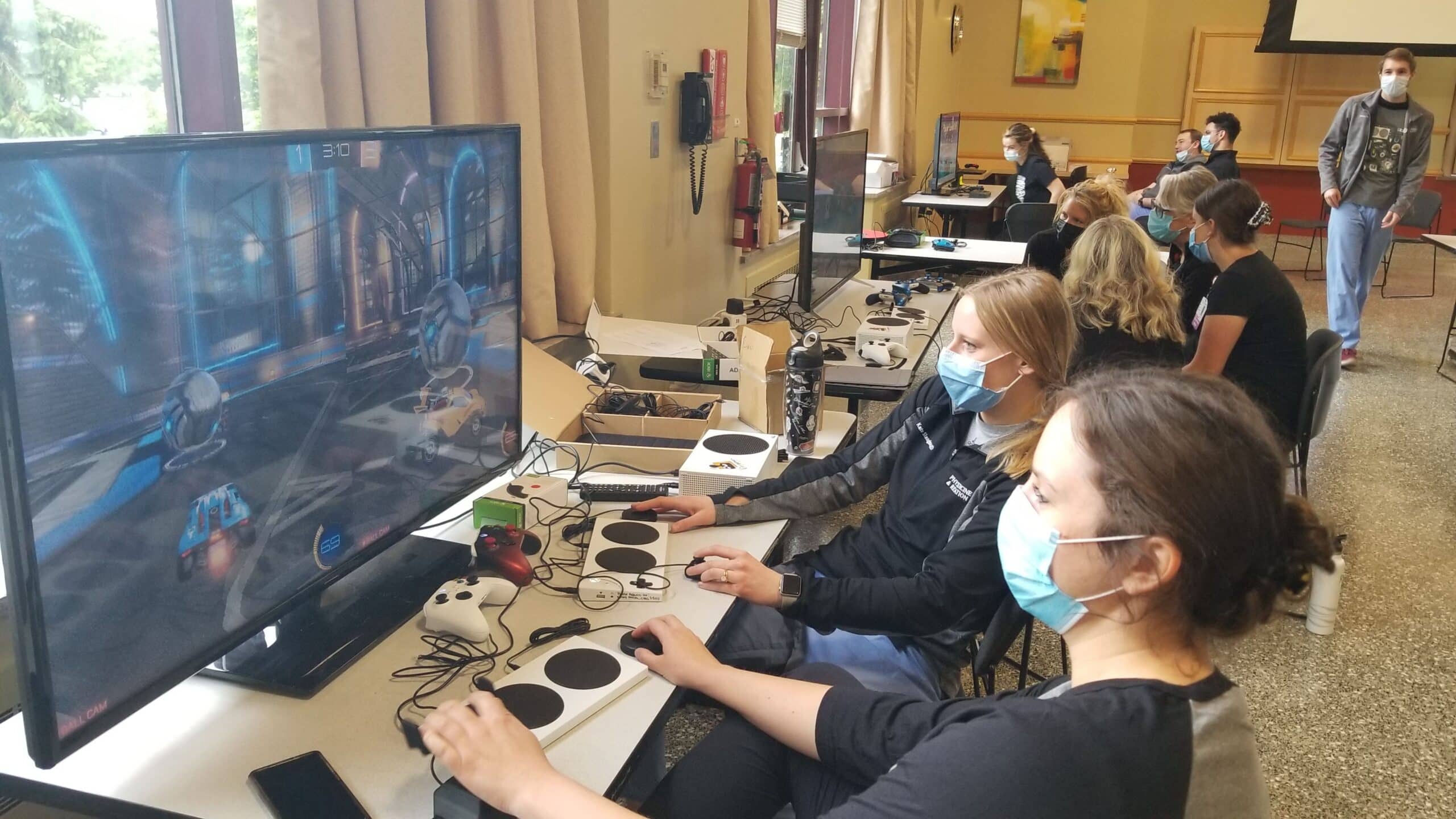 several people sitting in front of large monitors using various assistive gaming technologies such as the Xbox Adaptive Controller during a staff training session provided by the AbleGamers