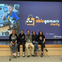 AbleGamers Peer Counseling staff members Jessie and Andy along with three Shepherd Center staff members sitting the edge of a stage, a large screen in back of them shows a slide with the AbleGamers logo from a presentation about the AbleGamers and Shepherd Center Partnership