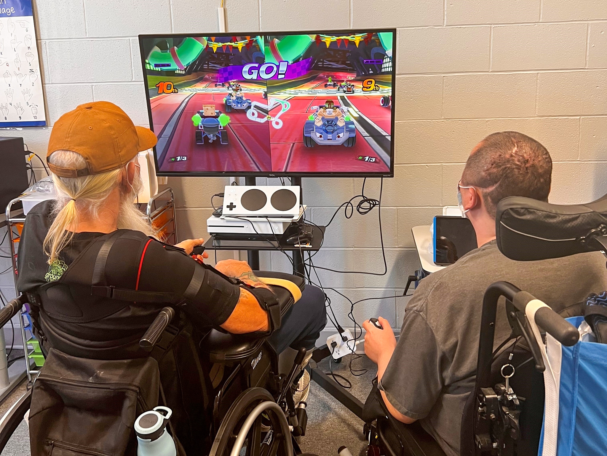 2 Men in wheelchairs, both looking at a screen playing a game. An XAC is on top of an xbox.