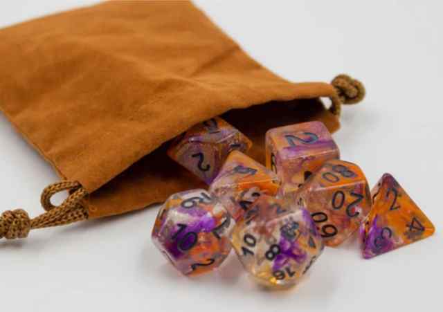 A bag of AbleGamers Dice showing 7 dice most commonly in Tabletop. The dice are clear with purple and orange swirls and the AbleGamers Joystick logo is placed on the 20 on the D20. 