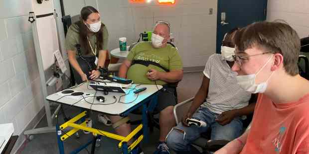three individuals playing video games, another person is standing near a neon Powered by AbleGamers sign. The photo was taken at Ability KC. Resources for People With Disabilities