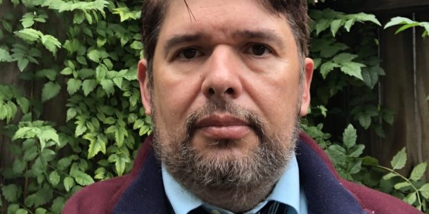 Photo of Roger Brooks. He is wearing a maroon fleece jacket with navy blue collar over a light blue dress shirt and navy and silver striped tie. He is standing in front of a wooden fence that is covered in leafy green vines.
