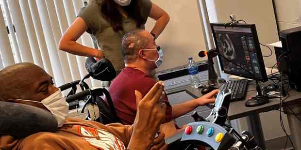 image provided by Ability KC one person standing in the back of the room two other persons using different adaptive gaming devices and controllers to play video games