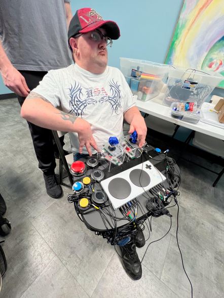 A photo provided by SEMCIL (SE MN Center for Independent Living) taken at one of their Adaptive Gaming Nights. A young man wearing a baseball cap and white t-shirt is playing a video game using several different buttons and switches mapped from an Xbox Adaptive Controller