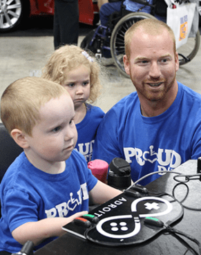 an image of three people, one man kneeling next to a young boy who is sitting in a chair and a young girl standing in the background
