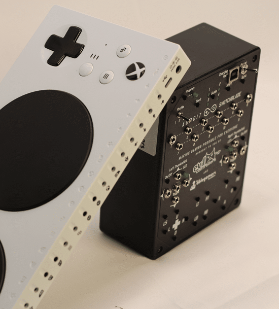 Adroit-and-MS-XAC An AbleGamers Adroit Switchblade, a black box with silver switch ports all over it. Leaning on the Adroit is a Microsoft XAC, a white candybar shapped device with ports on the back and a large black button.