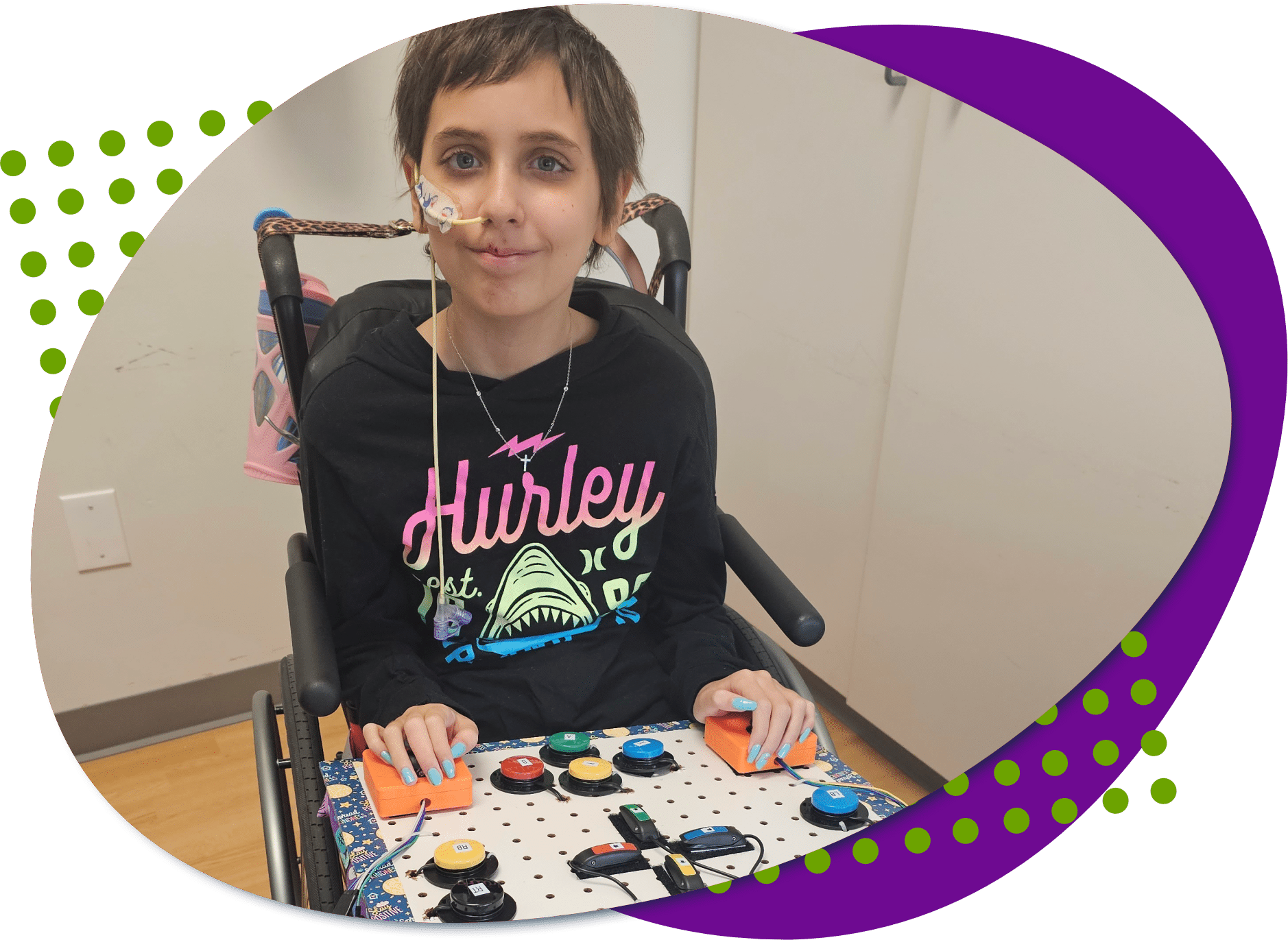 A photo of Bella, she has short brown hair and is wearing a black Hurley sweatshirt and she has blue painted nails. She is sitting and her new adaptive gaming set up is in front of her.
