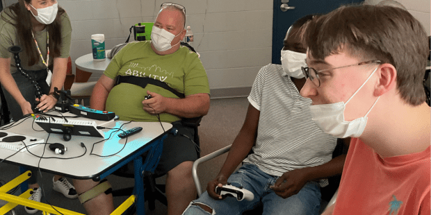 Three individuals playing a video game together and another indivdual standing in the background at Powered by AbleGamers partner Ability KC- Why Inclusive Gaming Matters