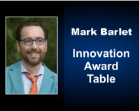 Mark Barlet head shot, he is wearing royal blue glasses, a teal suit jacket, blue dress shirt and AbleGamers orange tie. Next to Mark's photo is written Mark Barlet Innovation Award Table