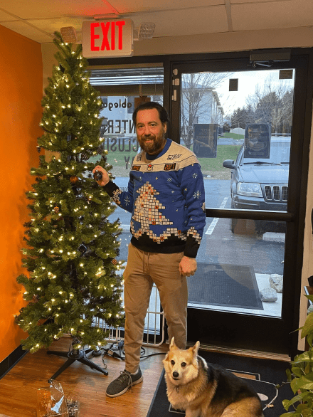 Founder Mark Barlet putting decorations on a christmas tree. Mark is a tall man with a beard who is wearing one of the Microsoft Ugly Sweaters. His dog, a corgi is by his feet