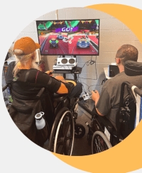 two men using wheelchairs are playing a racing video game. A large screen mounted on a cart in front of them.