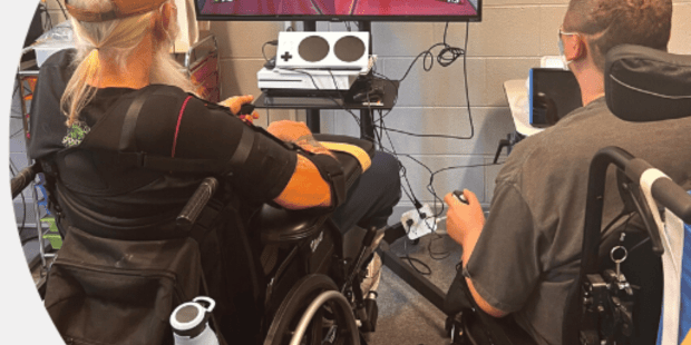 two men using wheelchairs are playing a racing video game. A large screen mounted on a cart in front of them.
