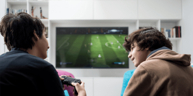 two teen boys playing a video game, one is wearing a brown hooded sweatshirt the other a black t-shirt. They are talking and smiling. The importance of connecting with others.