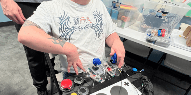 A photo provided by SEMCIL (SE MN Center for Independent Living) taken at one of their Adaptive Gaming Nights. A young man wearing a baseball cap and white t-shirt is playing a video game using several different buttons and switches mapped from an Xbox Adaptive Controller